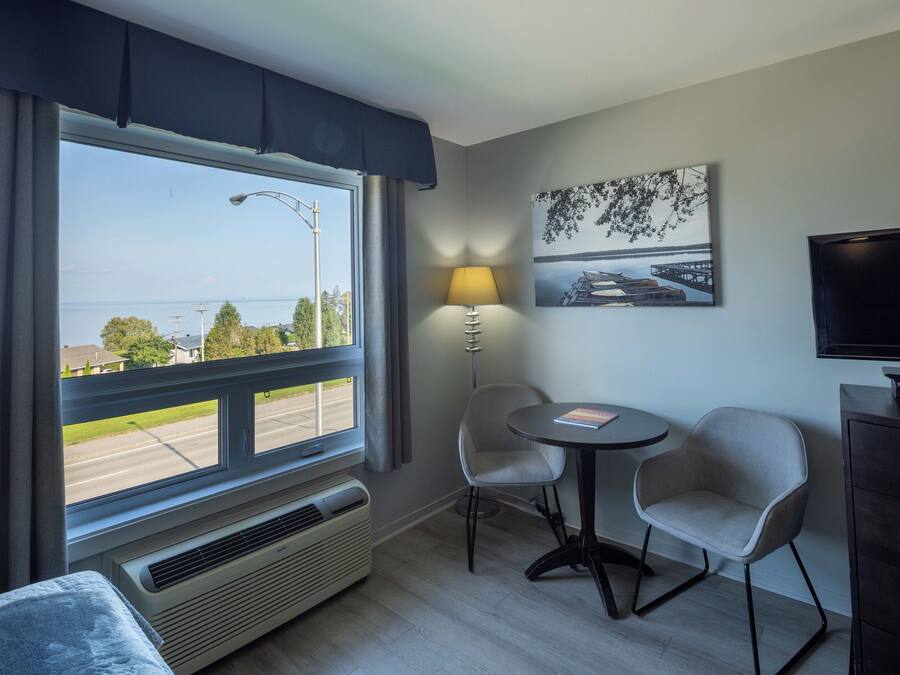 Habitación de hotel con un ventanal grande, una mesa con dos sillas, una lámpara, un televisor y un cuadro en la pared.