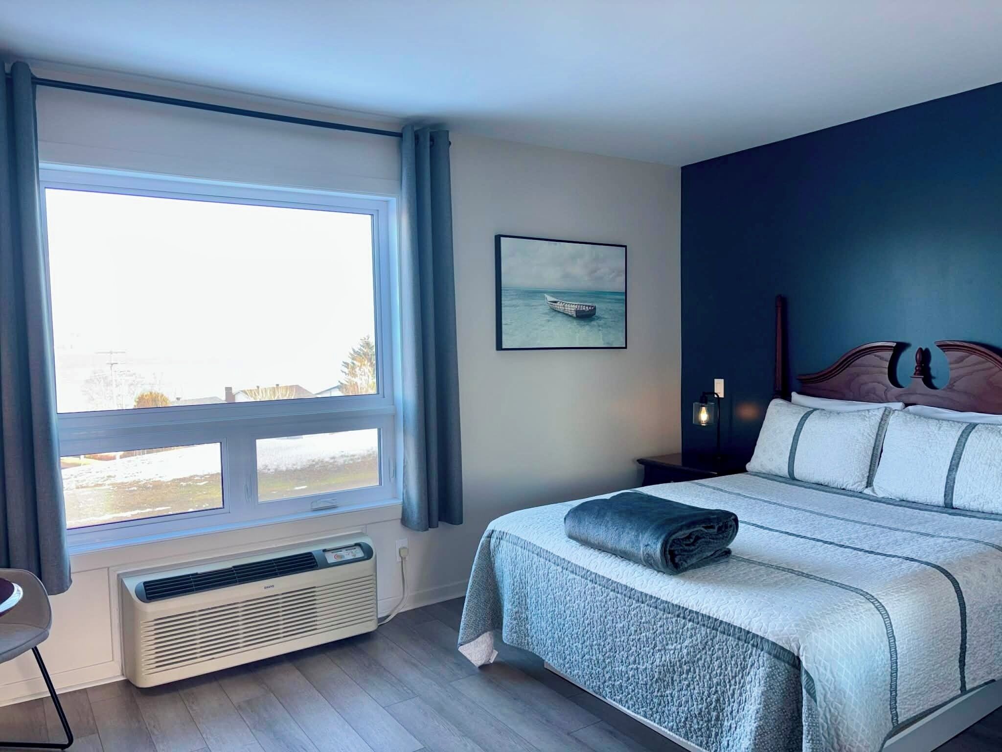A bedroom with a large window, a bed with a rolled-up blanket, a bedside table with a lamp, and a framed picture on the wall.