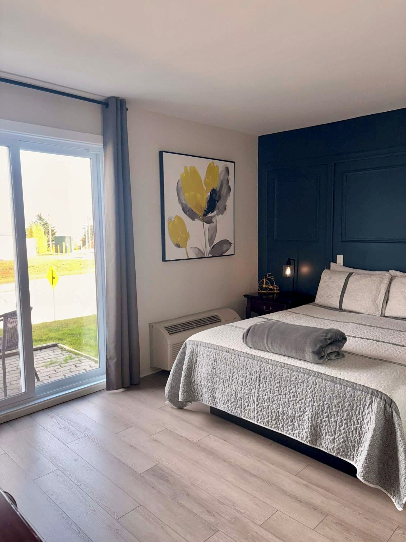 Un dormitorio con una cama grande, una mesita de noche, una obra de arte en la pared y un ventanal corredero que da a un patio.