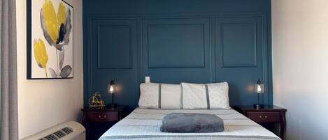 A bedroom with a bed, two bedside tables with lamps, a wall-mounted heater, and a framed floral artwork.