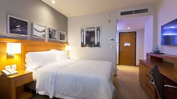 Habitación de hotel con cama, mesita de noche, lámpara y escritorio con silla. En la pared hay cuadros enmarcados y un televisor sobre el escritorio.