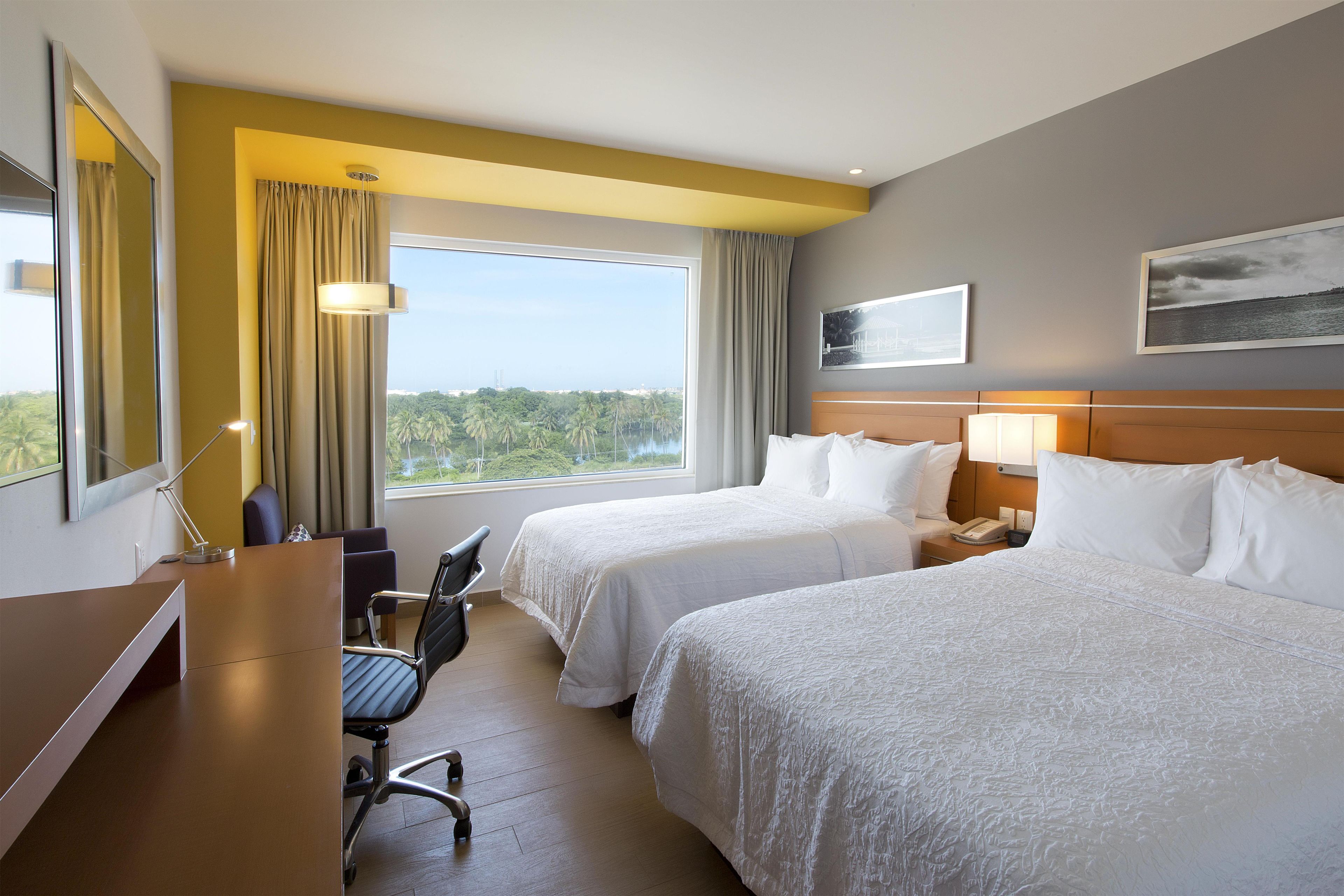 Een hotelkamer met twee bedden, een bureau, een stoel, een lamp en een raam met uitzicht op palmbomen.