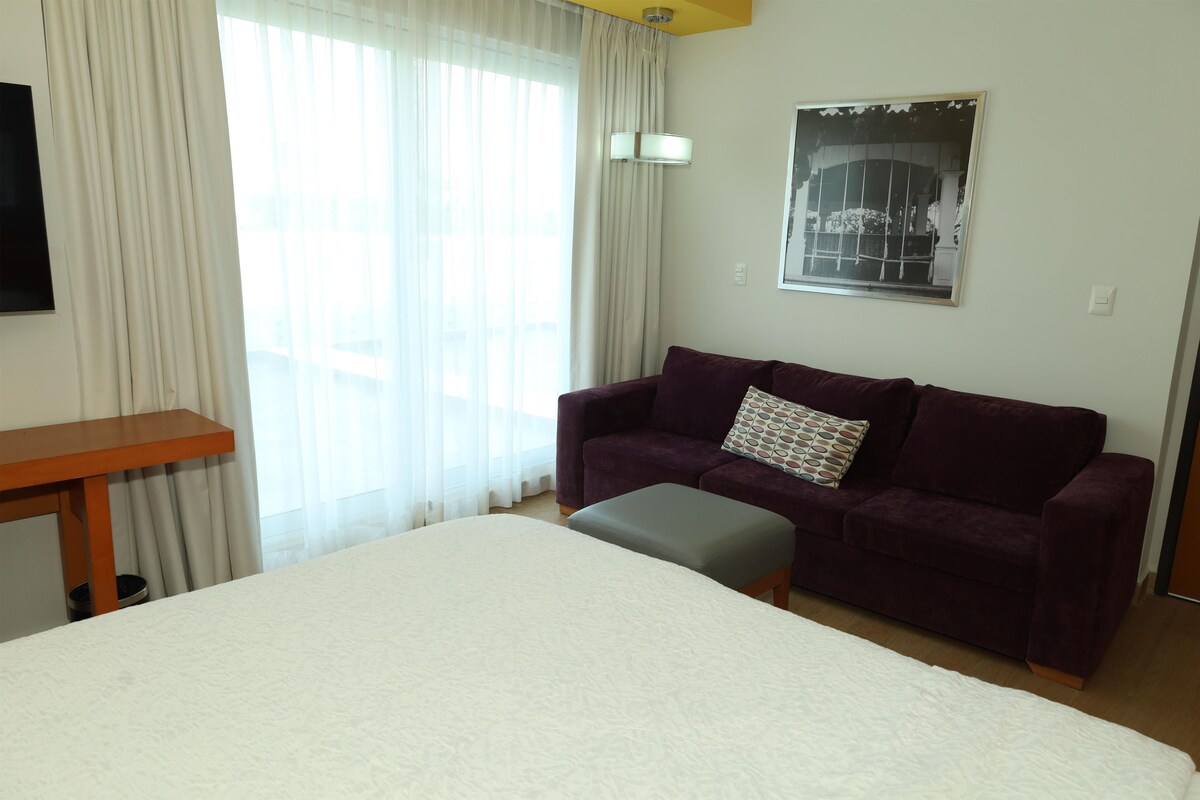 A hotel room with a bed, a purple sofa, a small ottoman, a wooden side table, and a window with sheer curtains.