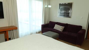 Habitación de hotel con cama, sofá morado, pequeño otoman, mesita de madera y ventana con cortinas translúcidas.
