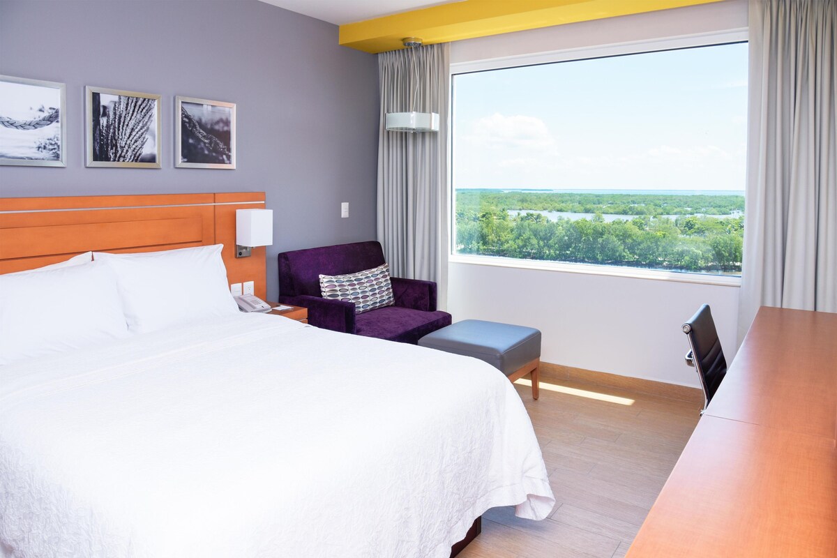 A hotel room with a bed, a chair, a table, and a large window offering a view of a landscape.