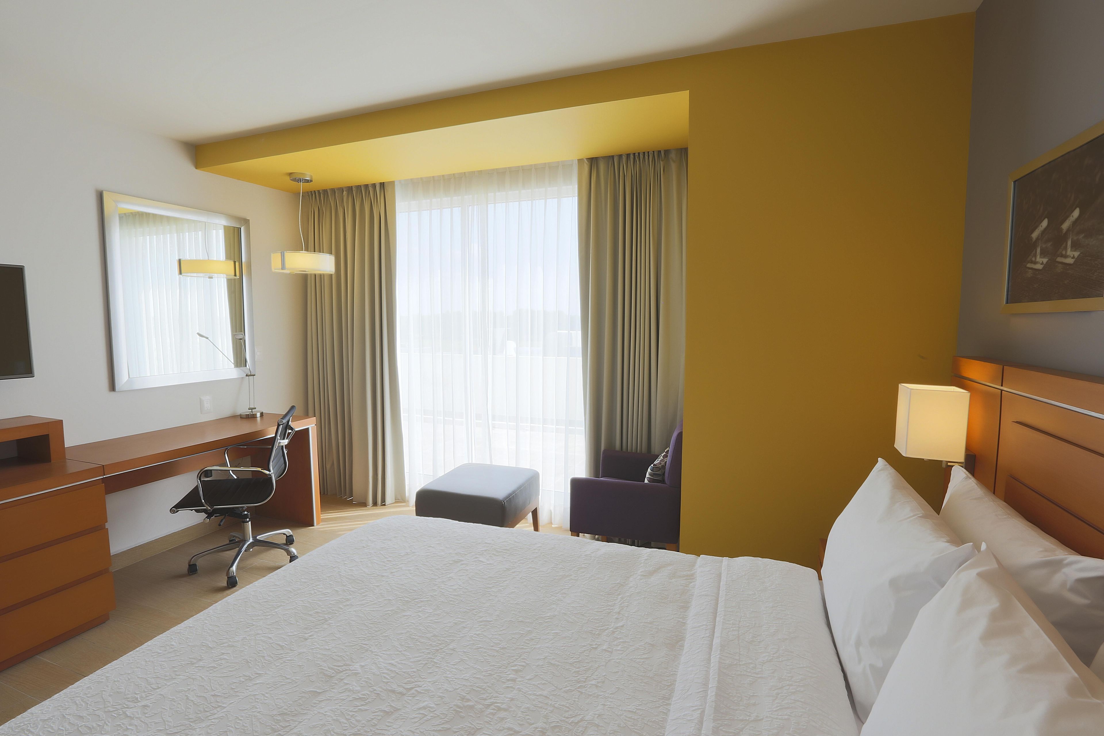 A hotel room with a bed, a desk, a chair, a lamp, and a window with curtains.