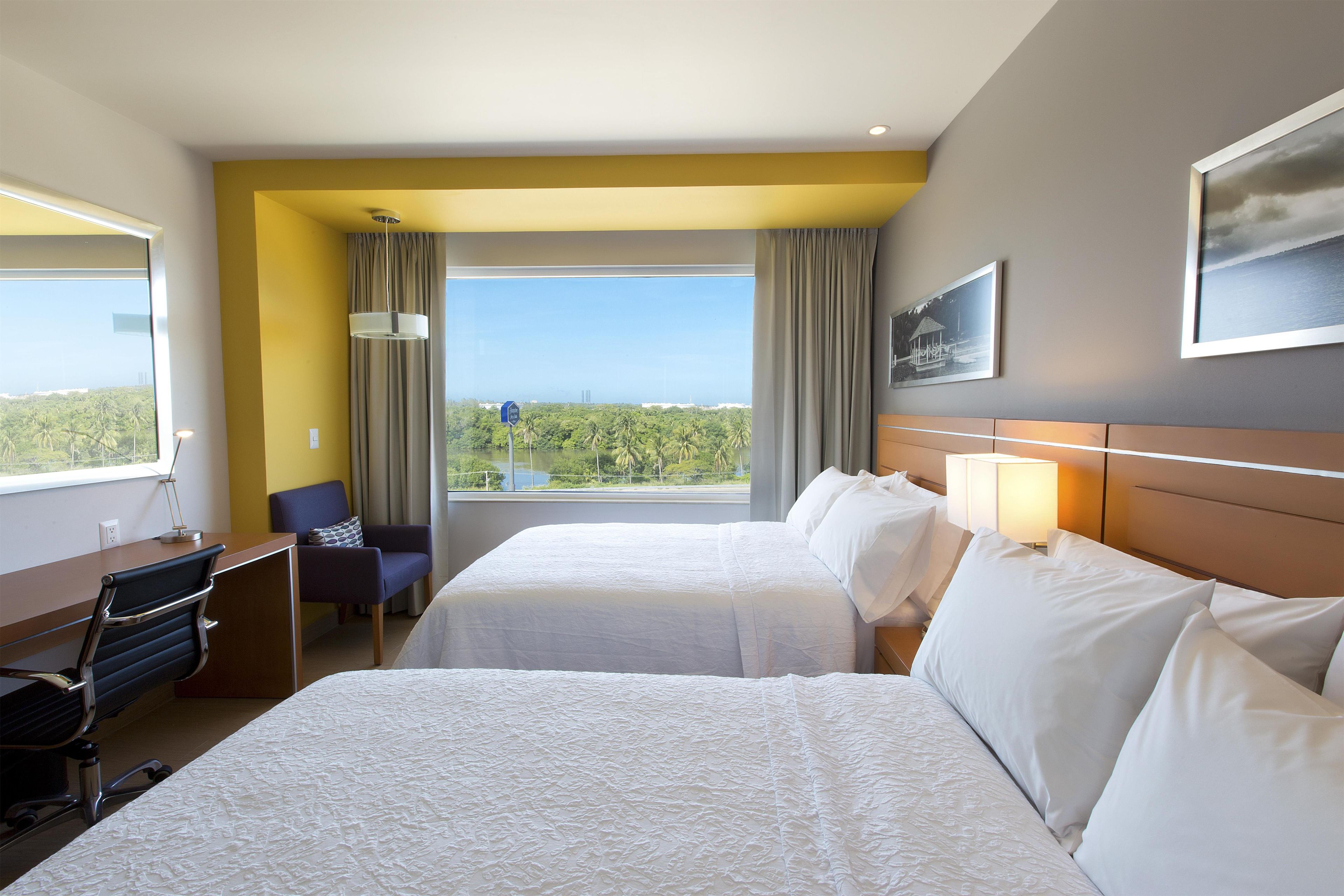 Habitación de hotel con dos camas, un escritorio, una silla, vistas al exterior y un cuadro en la pared.