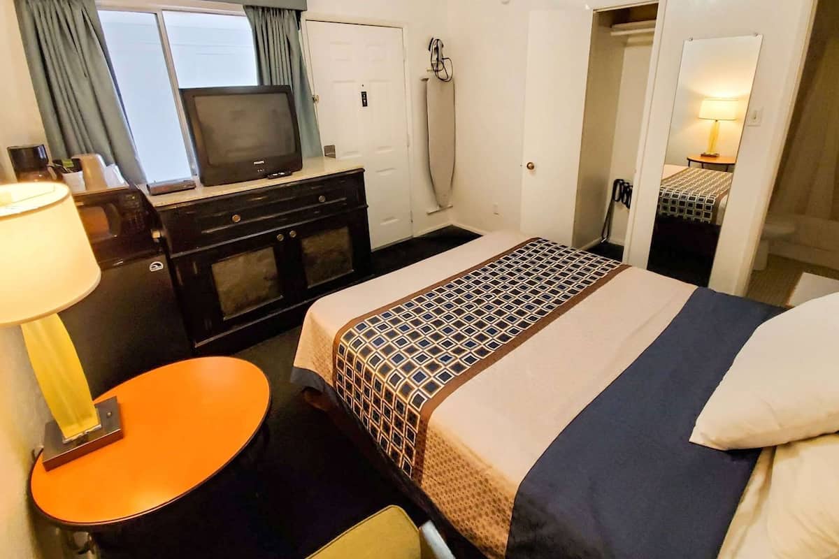 Habitación de hotel con cama, escritorio con lámpara, televisor, armario y baño visible a través de una puerta abierta.
