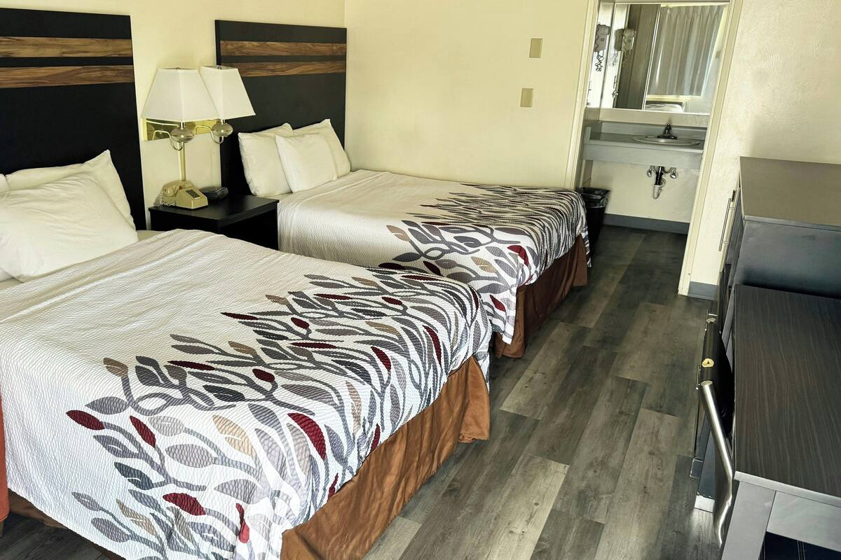 Habitación de hotel con dos camas, un escritorio, una silla, una mesita de noche con lámparas y un baño visible a través de una puerta abierta.