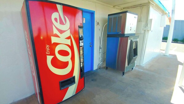 Una máquina expendedora roja de Coca-Cola junto a una puerta azul, con un pequeño equipo de aire acondicionado al fondo.