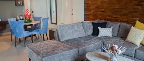 Una sala de estar moderna con un sofá seccional gris, un reposapiés a juego y una mesa de centro redonda. Hay una zona de comedor con sillas azules y una mesa, y una cocina al fondo.