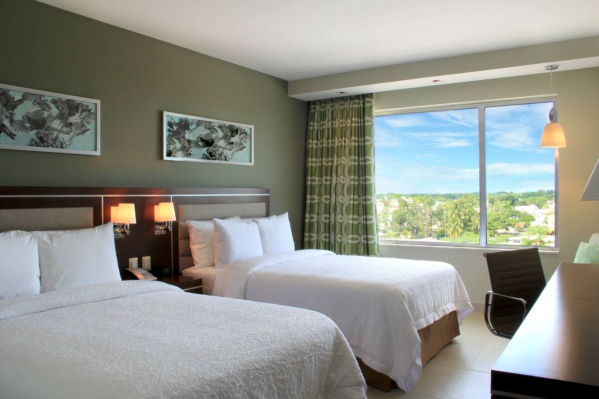 Une chambre d’hôtel avec deux lits, un bureau, une chaise et une grande fenêtre donnant sur un beau paysage.