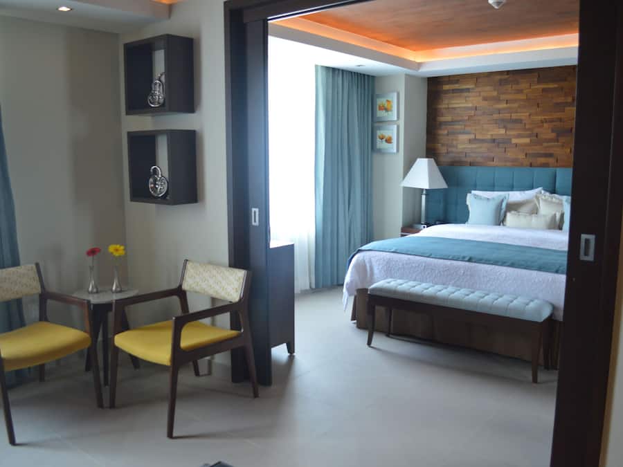 A hotel room with a bed, a desk with two chairs, a lamp, and a wall with framed pictures.