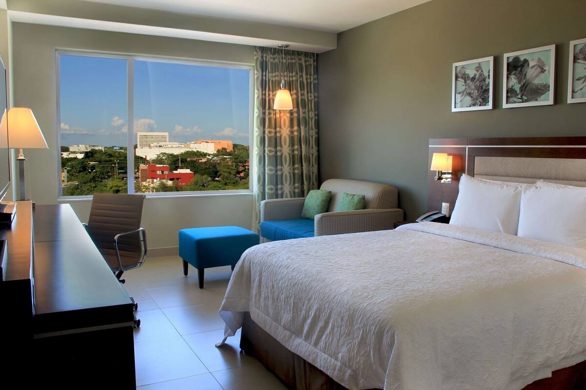 Une chambre d’hôtel avec un grand lit, un bureau, une chaise, un petit pouf et une vue sur le paysage urbain par la fenêtre.