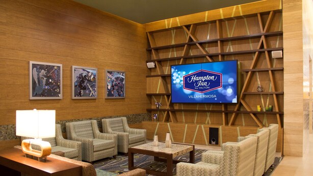 Hotellin aula, jossa on suuri näyttö, jolla esitetään tekstiä ”Hampton Inn by Hilton”, kolme kehystettyä taideteosta sekä oleskelualue, jossa on kuvioituja sohvia ja sohvapöytä.