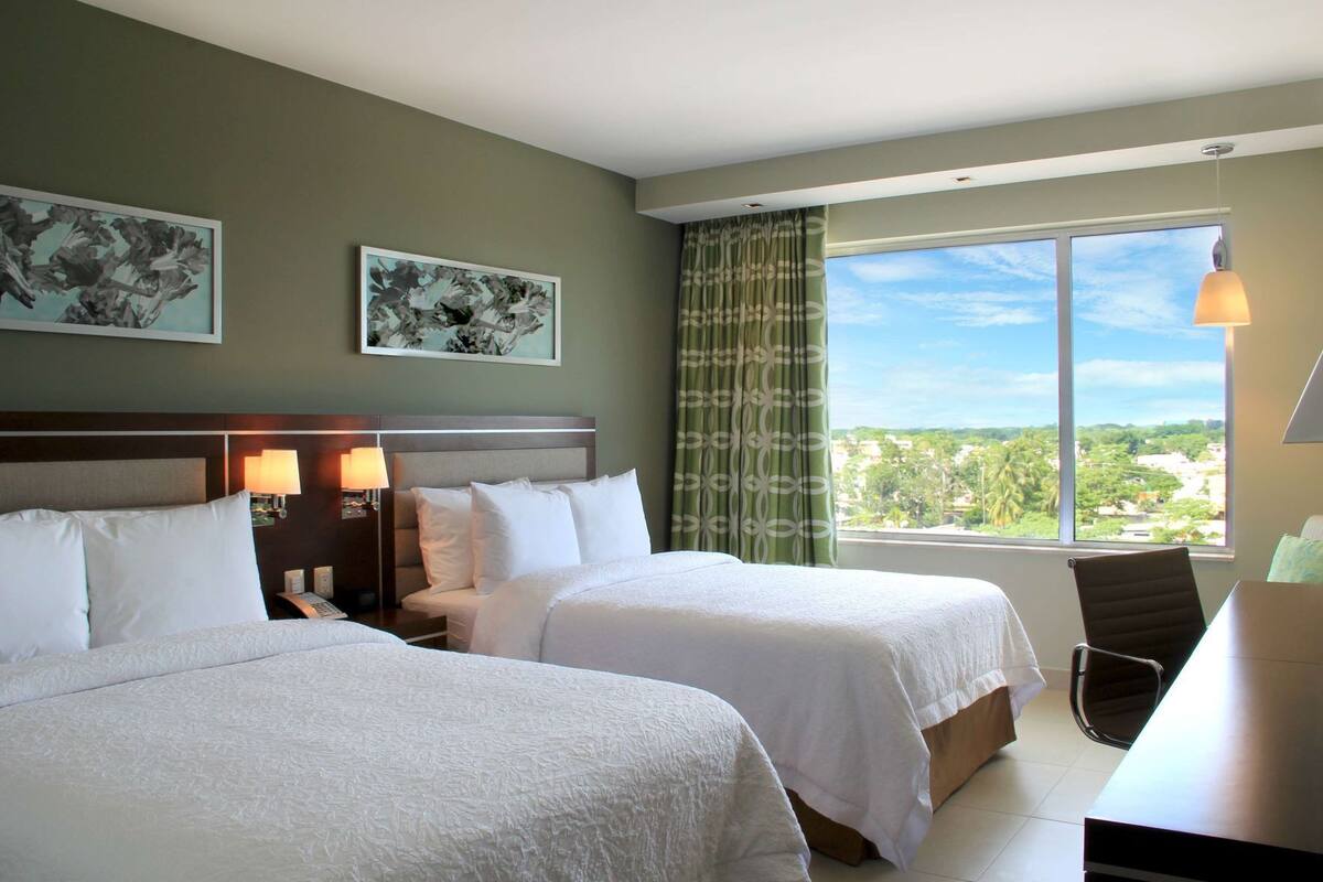 Une chambre d’hôtel avec deux lits, un bureau, une chaise, une fenêtre donnant sur un beau paysage et des œuvres d’art accrochées aux murs.