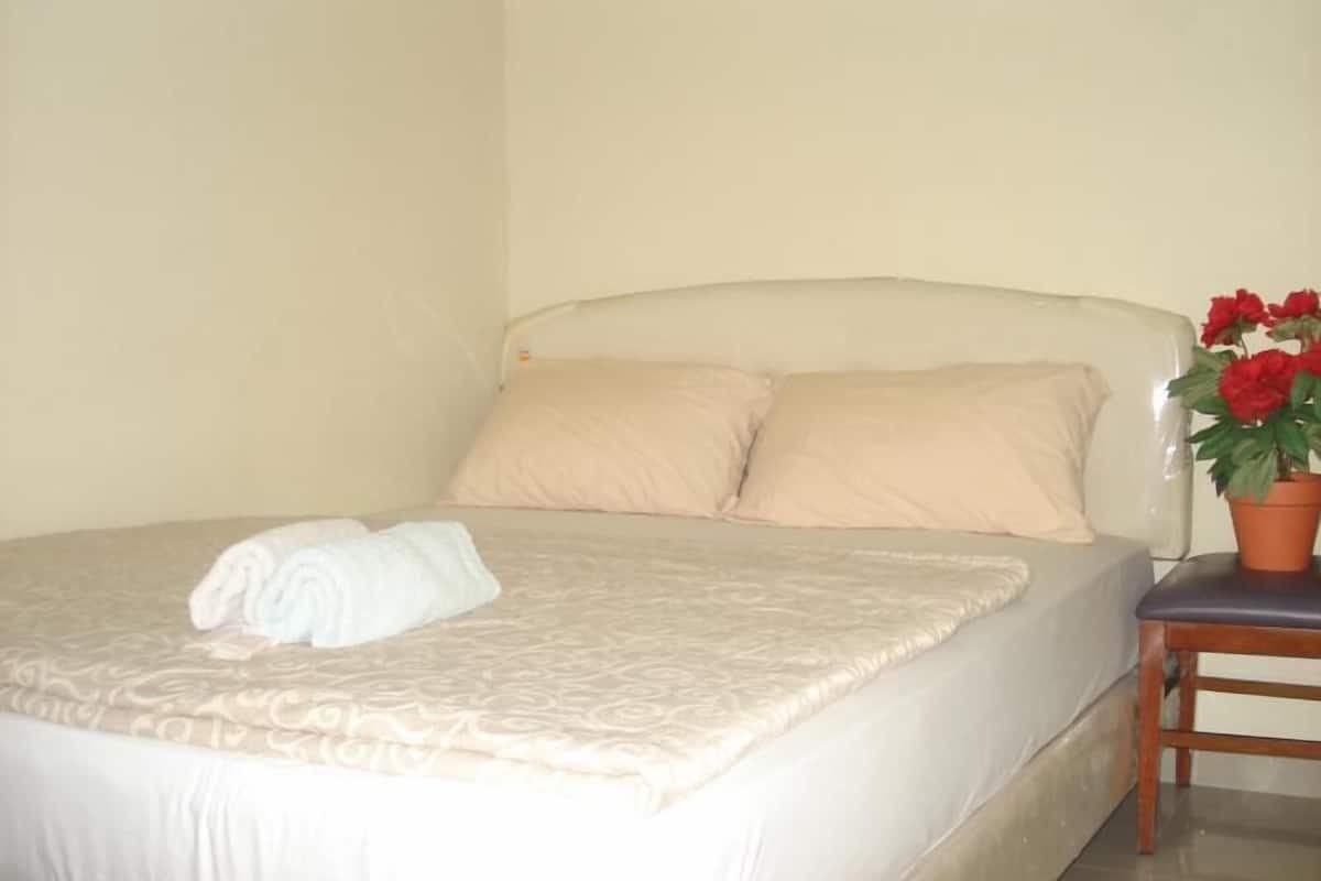 Un letto con copriletto beige, due cuscini, un asciugamano bianco arrotolato e una pianta in vaso con fiori rossi su un comodino in legno.