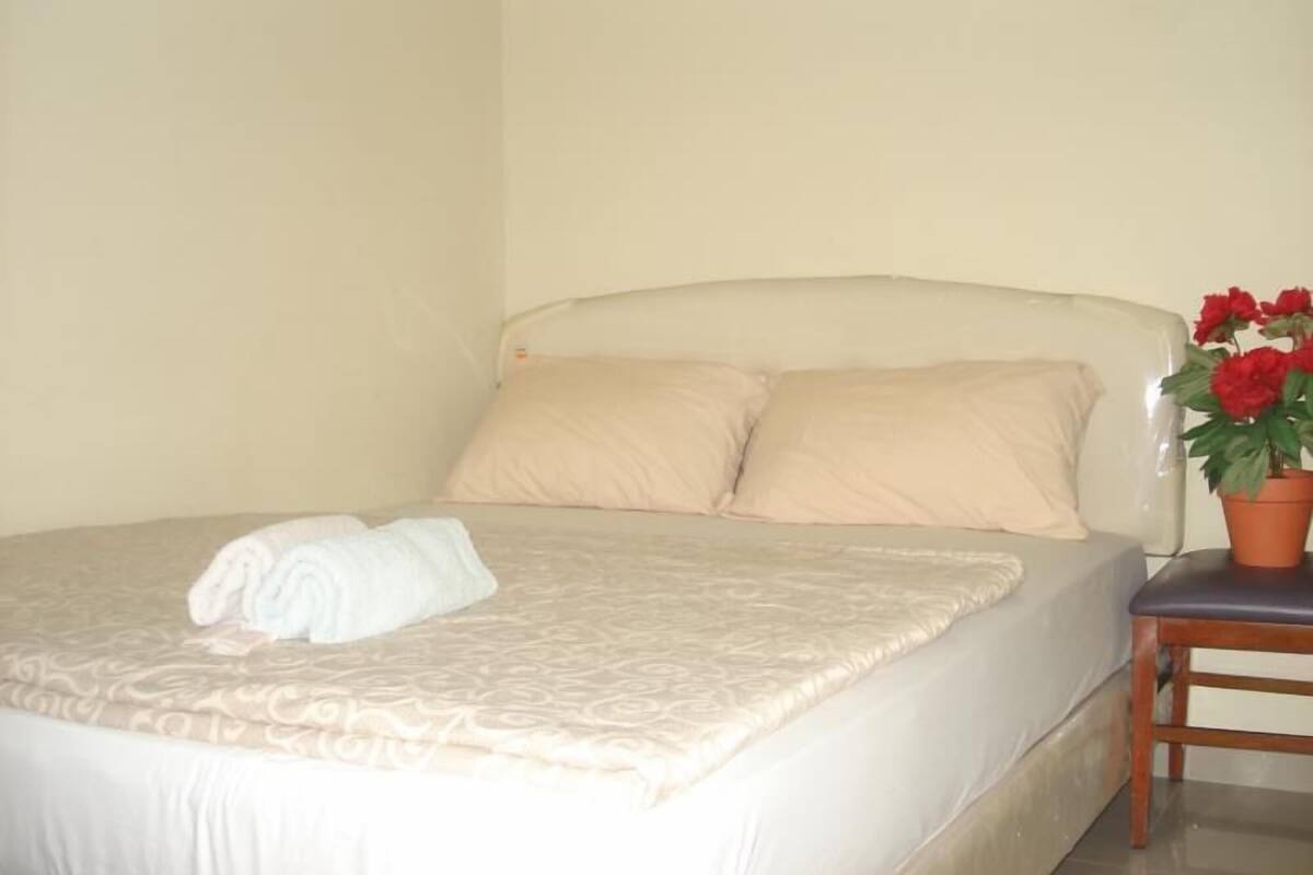 Un letto con copriletto beige, due cuscini, un asciugamano bianco arrotolato e una pianta in vaso con fiori rossi su un comodino in legno.
