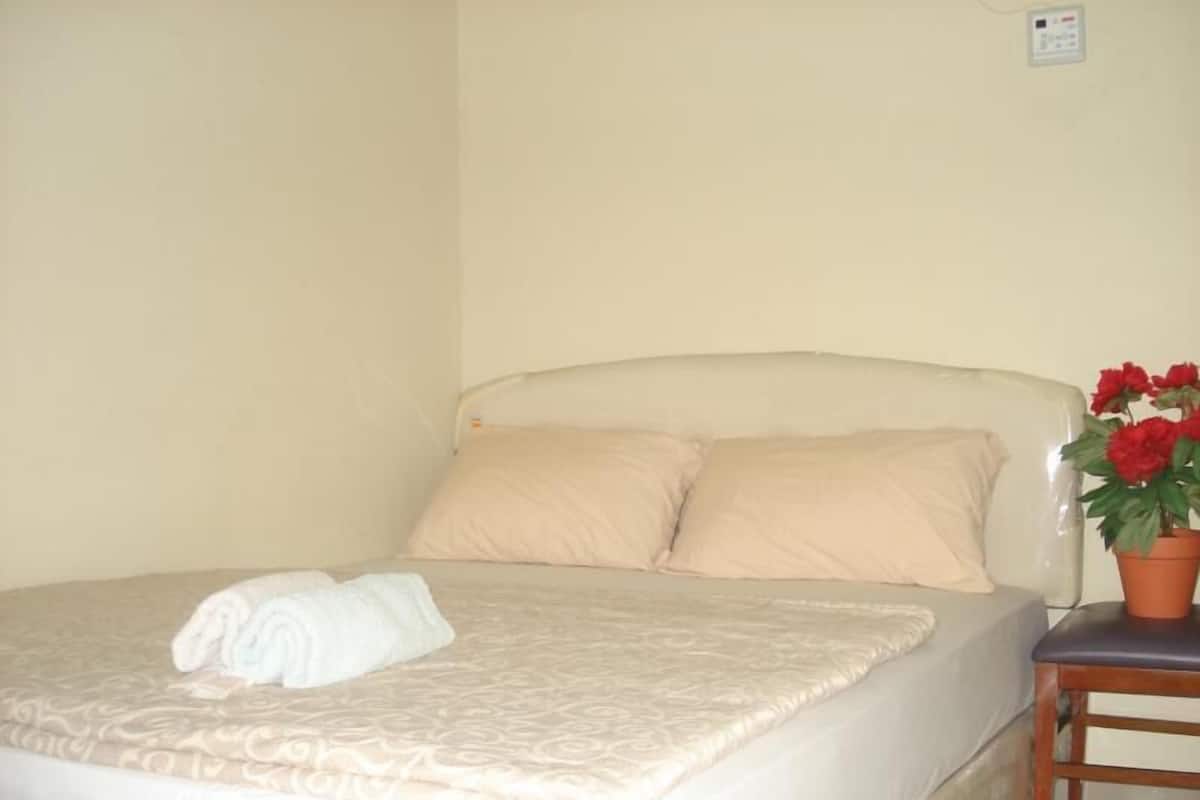 Un letto rifatto con cura, con copriletto beige e cuscini, un asciugamano bianco arrotolato sul letto e un vaso con fiori rossi su un comodino in legno accanto al letto.