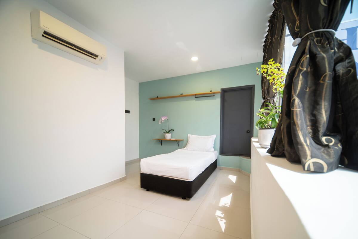 Une chambre à coucher comprenant un lit, un appareil de climatisation, une porte, une étagère avec un pot de plante et une fenêtre avec des rideaux.