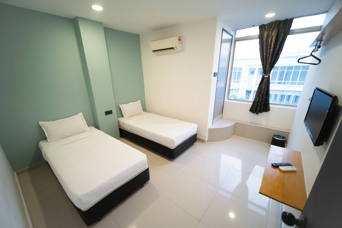 Une chambre d’hôtel avec deux lits simples, une petite table avec un téléphone, un téléviseur fixé au mur et une fenêtre avec des rideaux.