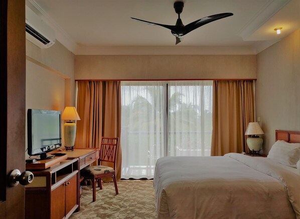 A hotel room with a bed, a desk with a computer, a chair, a ceiling fan, and a window with curtains.