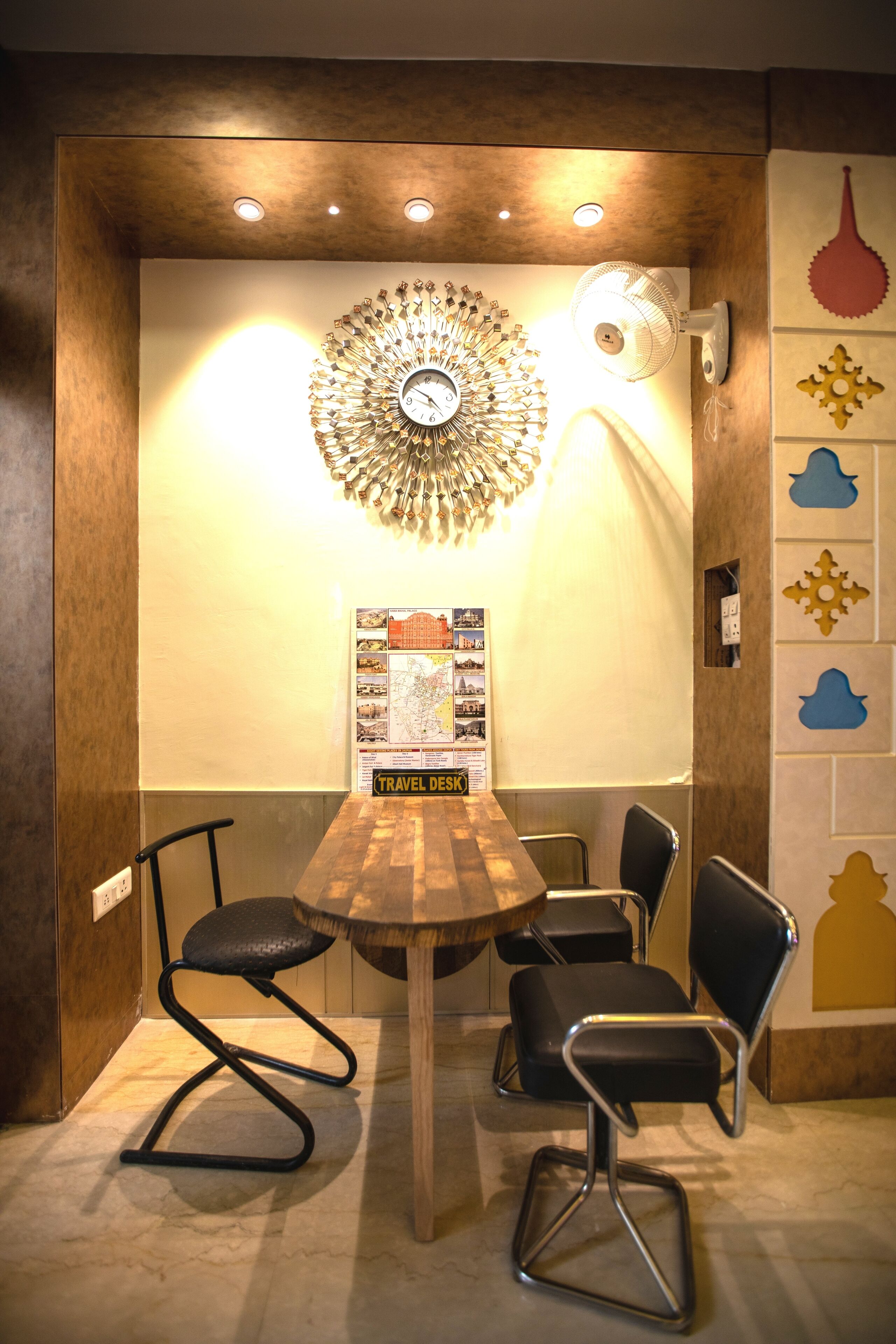 A small meeting room with a wooden table, four black chairs, a decorative wall clock, a wall-mounted fan, and a travel desk with a map.