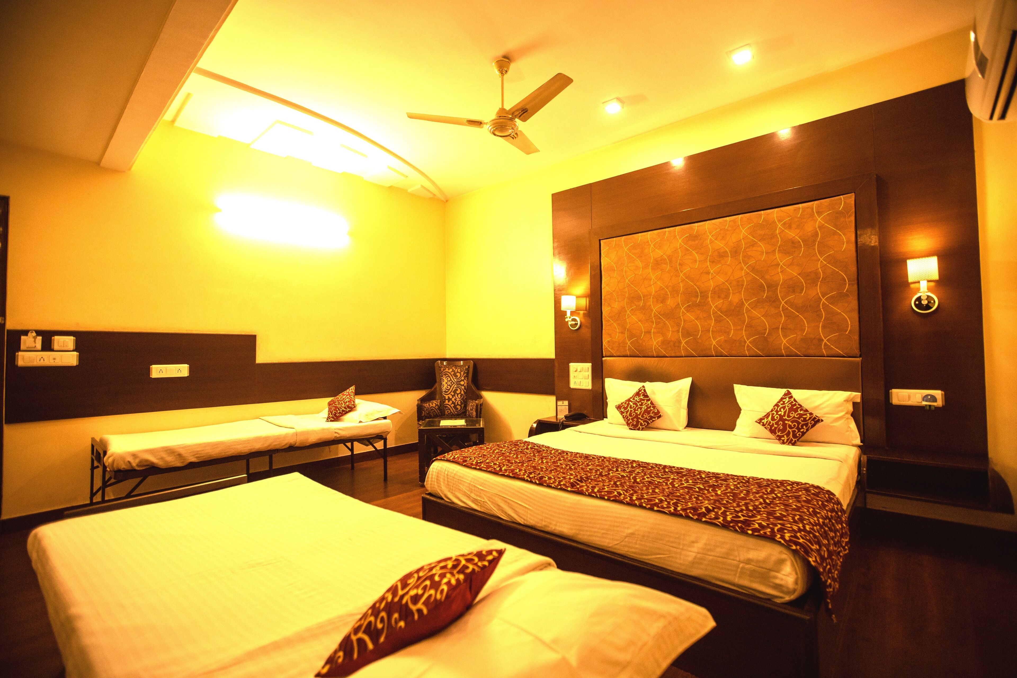 A hotel room with two single beds, a double bed, a ceiling fan, bedside lamps, and a patterned headboard.