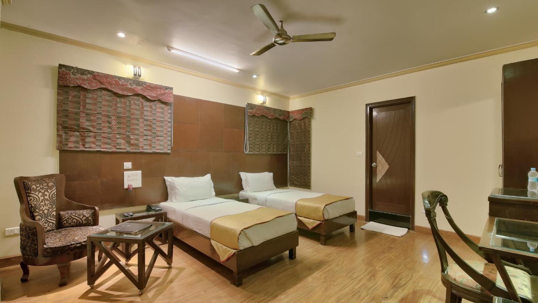 A hotel room with two single beds, a wooden chair, a small table, a ceiling fan, and a door.