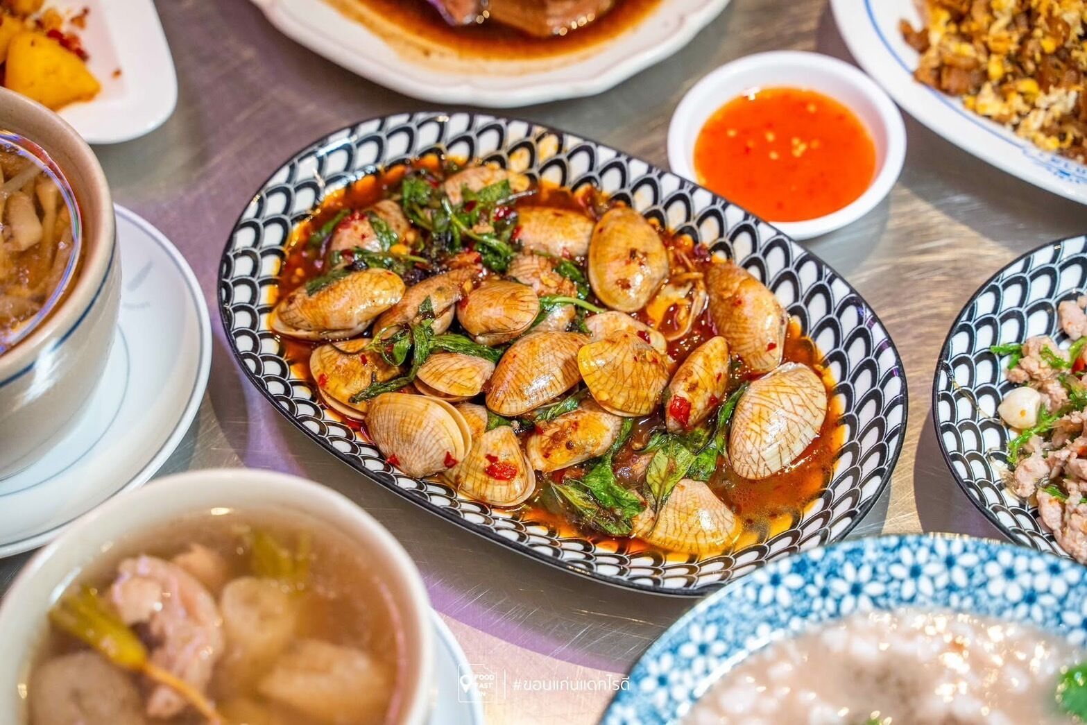 A variety of dishes on a table, including a bowl of clams in a spicy sauce, a bowl of soup, and a plate of stir-fried meat.