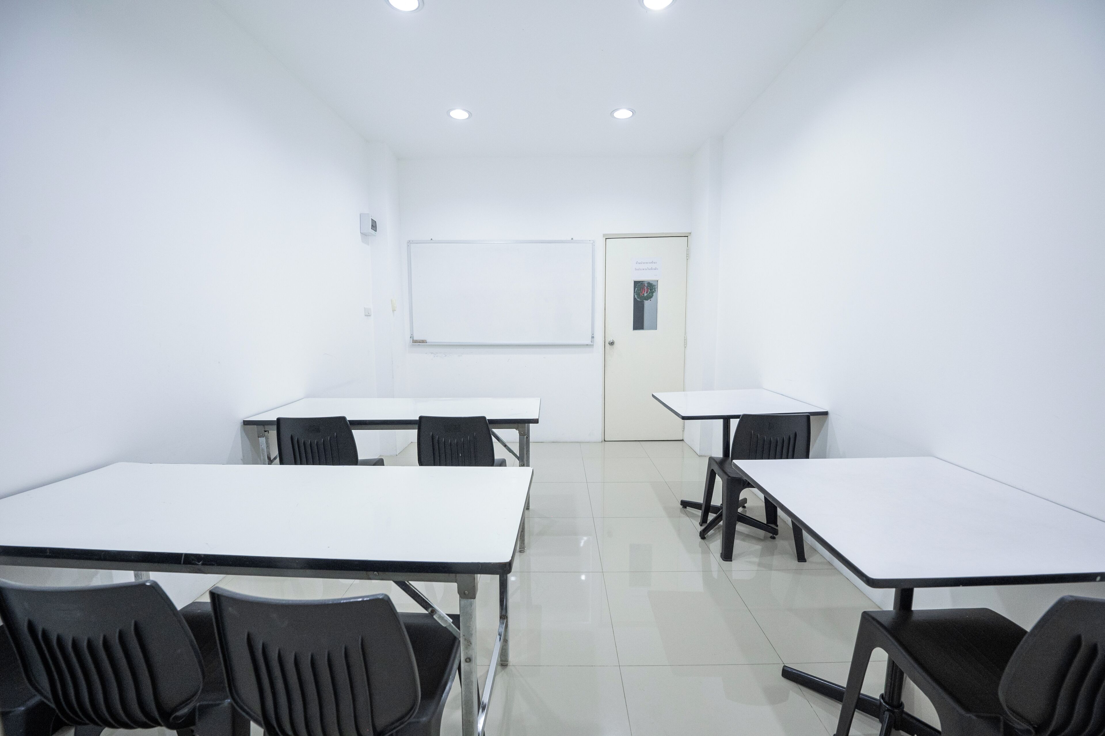 A conference room with white tables and black chairs, a whiteboard, and a door with a sign.