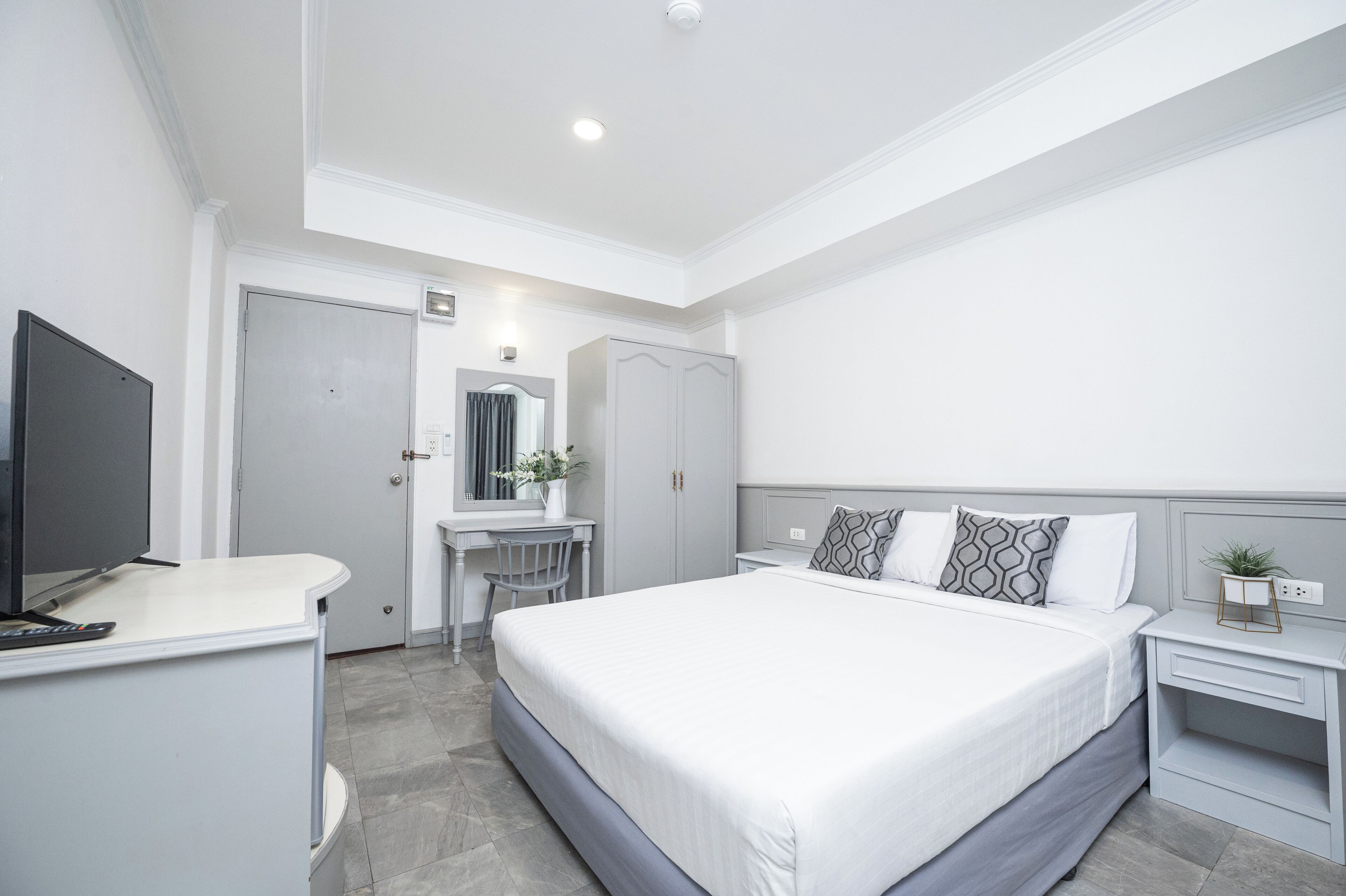 A modern hotel room with a large bed, a desk with a TV, a vanity with a mirror, and a wardrobe.