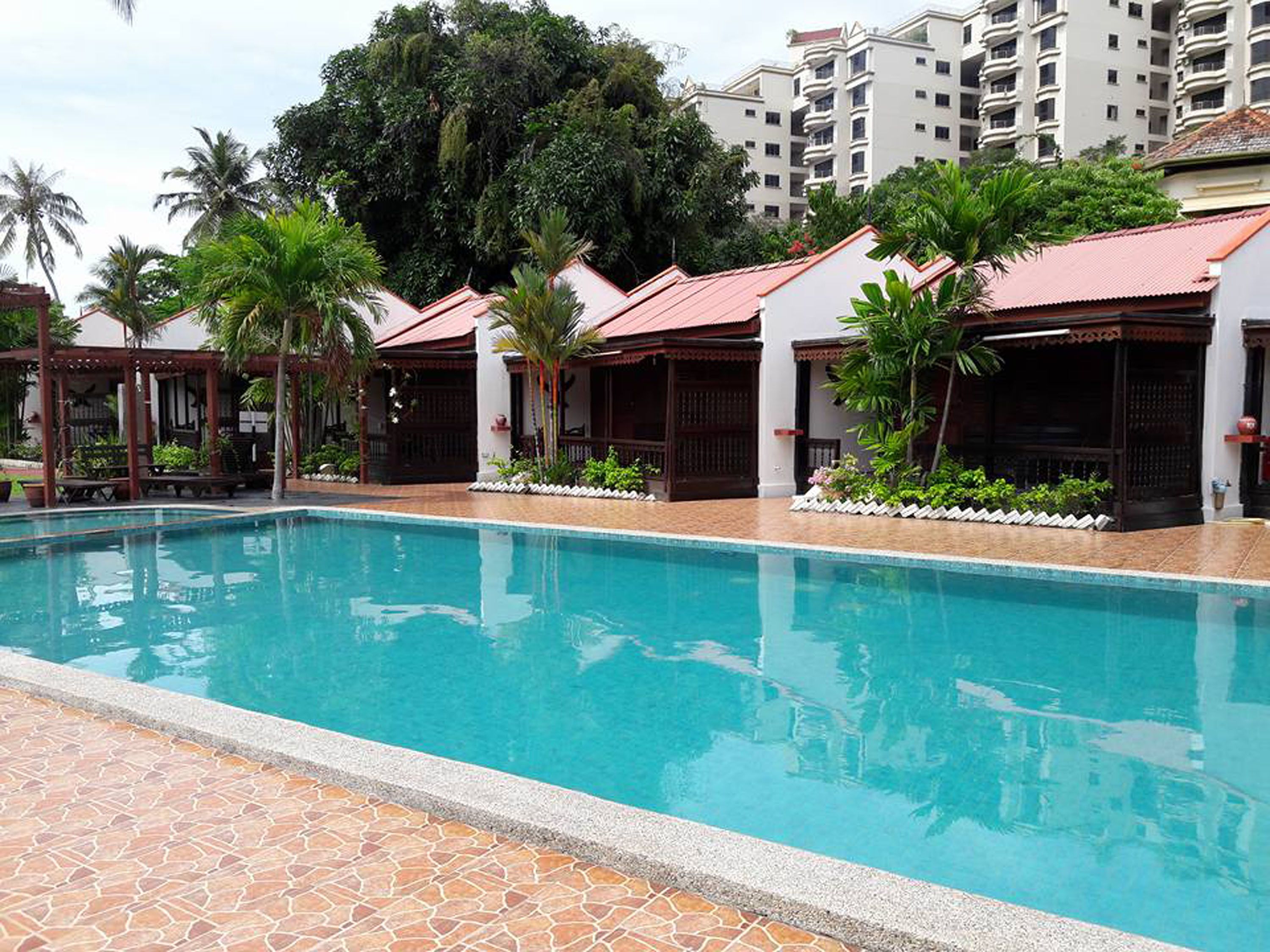 A swimming pool with tiled edges, surrounded by palm trees and a building with a red roof.