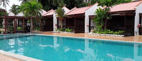 A swimming pool with tiled edges, surrounded by palm trees and a building with a red roof.