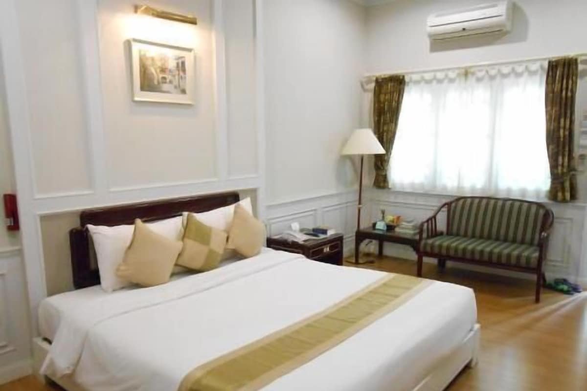 A hotel room with a large bed, a bedside table, a chair, a lamp, a picture on the wall, and an air conditioning unit.