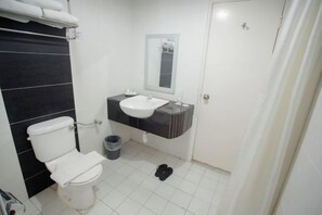 Une salle de bain avec des toilettes, un lavabo et un miroir. On y trouve une poubelle et une paire de chaussures sur le sol.