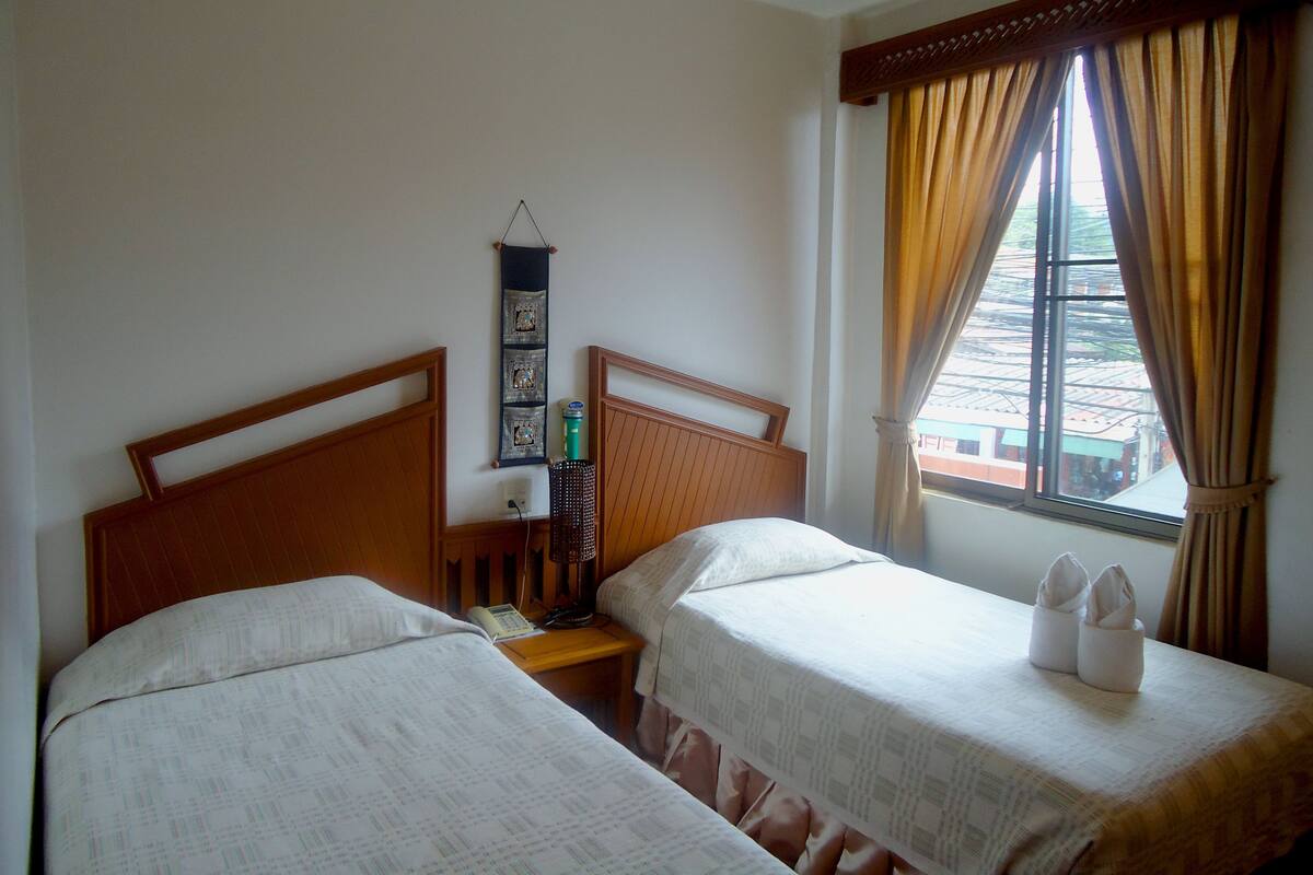 Een hotelkamer met twee eenpersoonsbedden, een houten hoofdeinde, een klein nachtkastje met een lamp, een decoratie aan de muur en een raam met gordijnen.