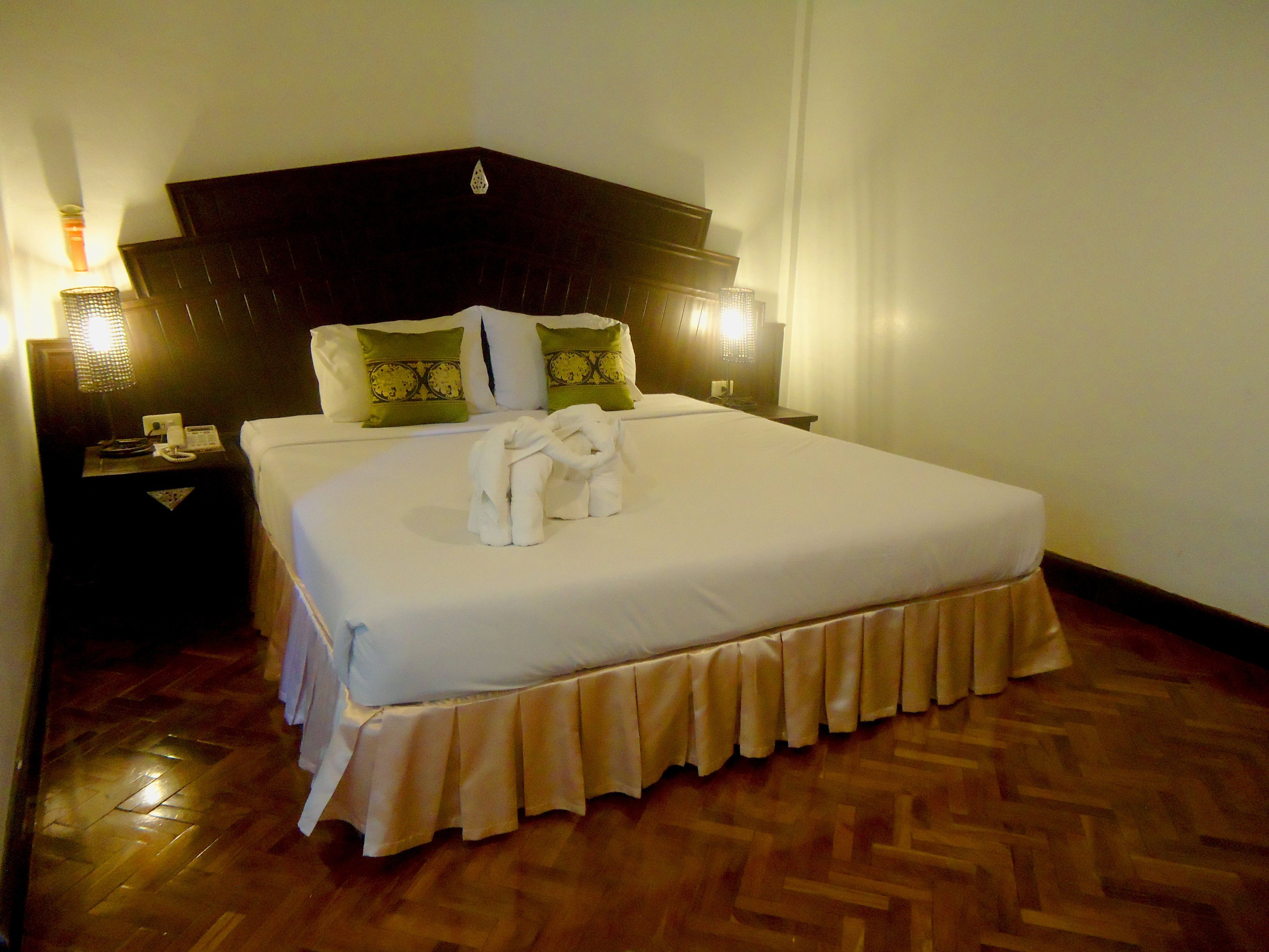 Una cama bien hecha con una colcha blanca y un arreglo decorativo de toallas. Hay dos almohadas con un estampado verde y dorado. La habitación tiene piso de madera y un cabecero oscuro.
