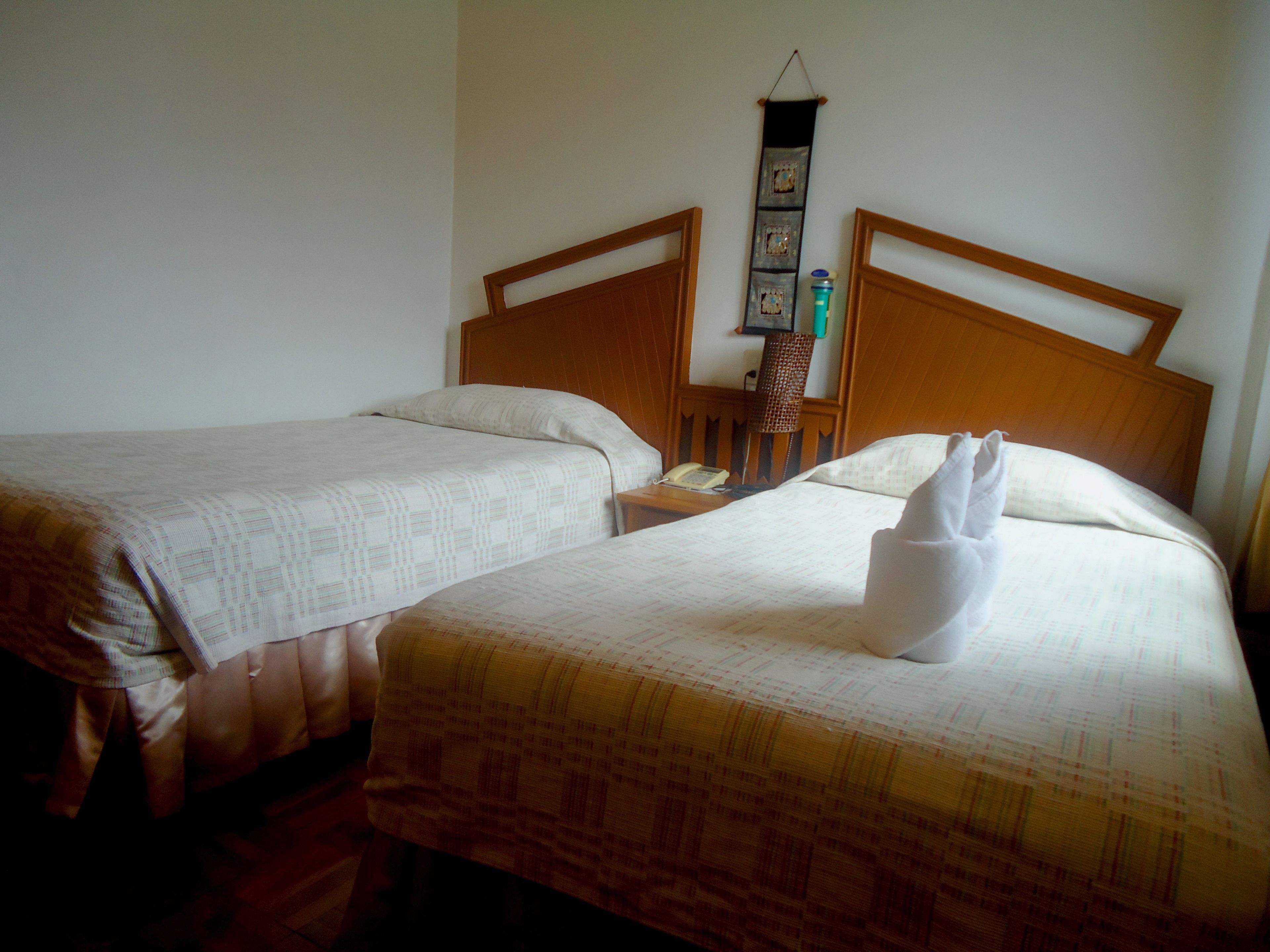 Habitación de hotel con dos camas, cada una con cabecero de madera, una mesita de noche con lámpara y teléfono en la pared.