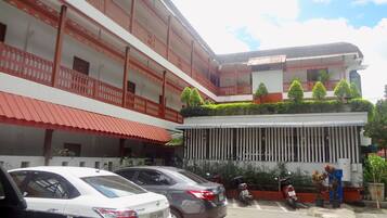 A multi-story building with a red and white facade, balconies, and a parking area with cars and motorcycles.