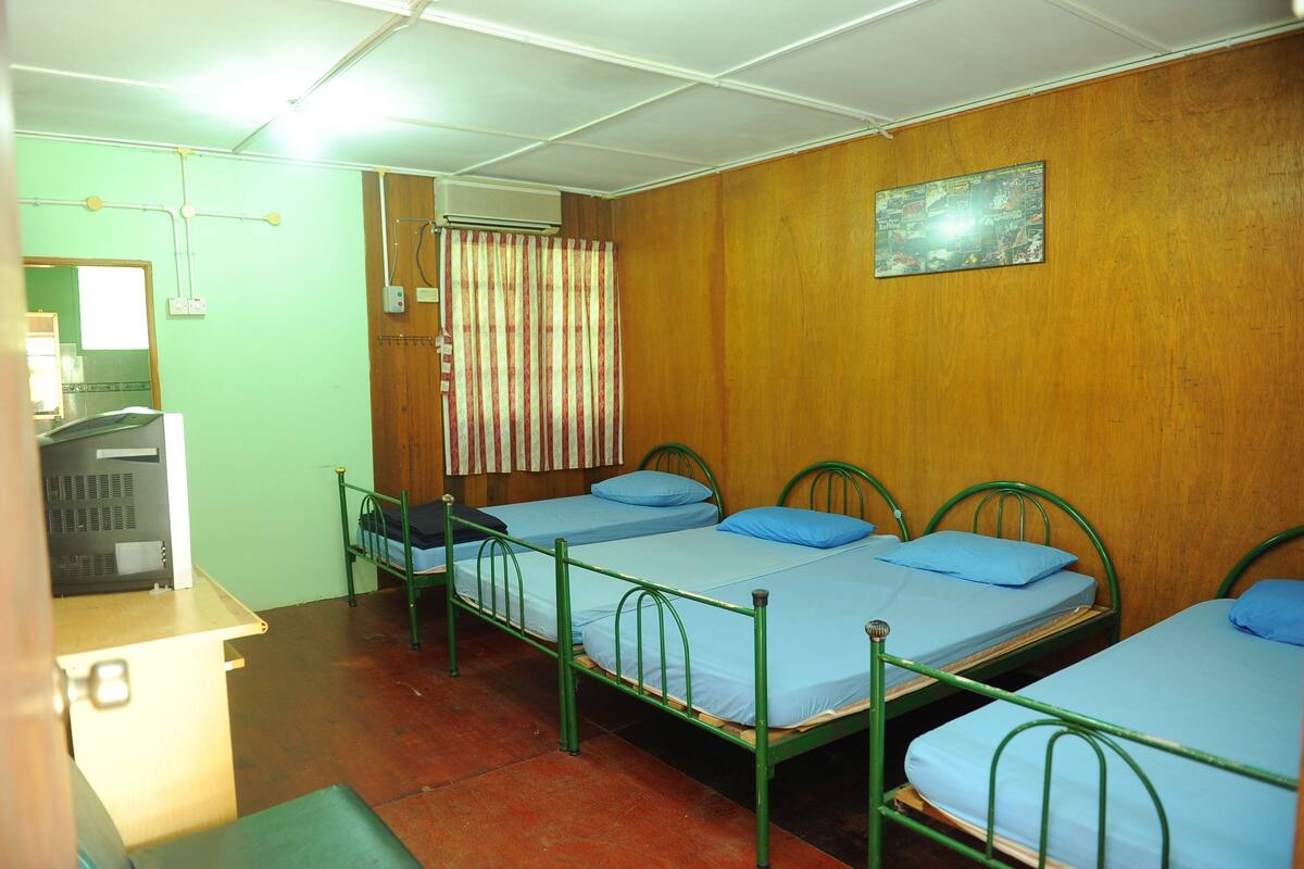 Une chambre de dortoir avec trois lits superposés en métal, un mur en bois, un lit simple avec un matelas bleu, un petit bureau et une fenêtre avec des rideaux.