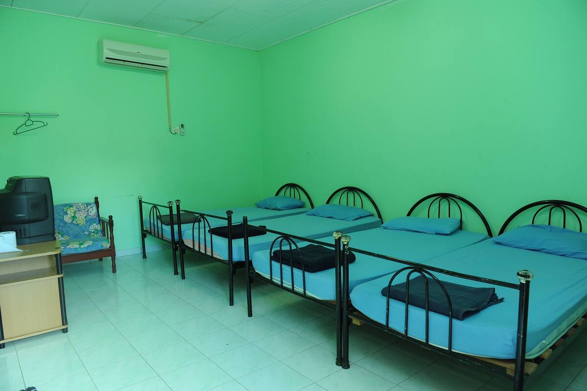Une chambre de dortoir avec quatre lits en métal, un téléviseur posé sur une commode en bois et un climatiseur fixé au mur.