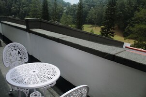 Uma varanda com mesa e cadeira de metal branco, um parapeito de pedra e vista para uma área arborizada.