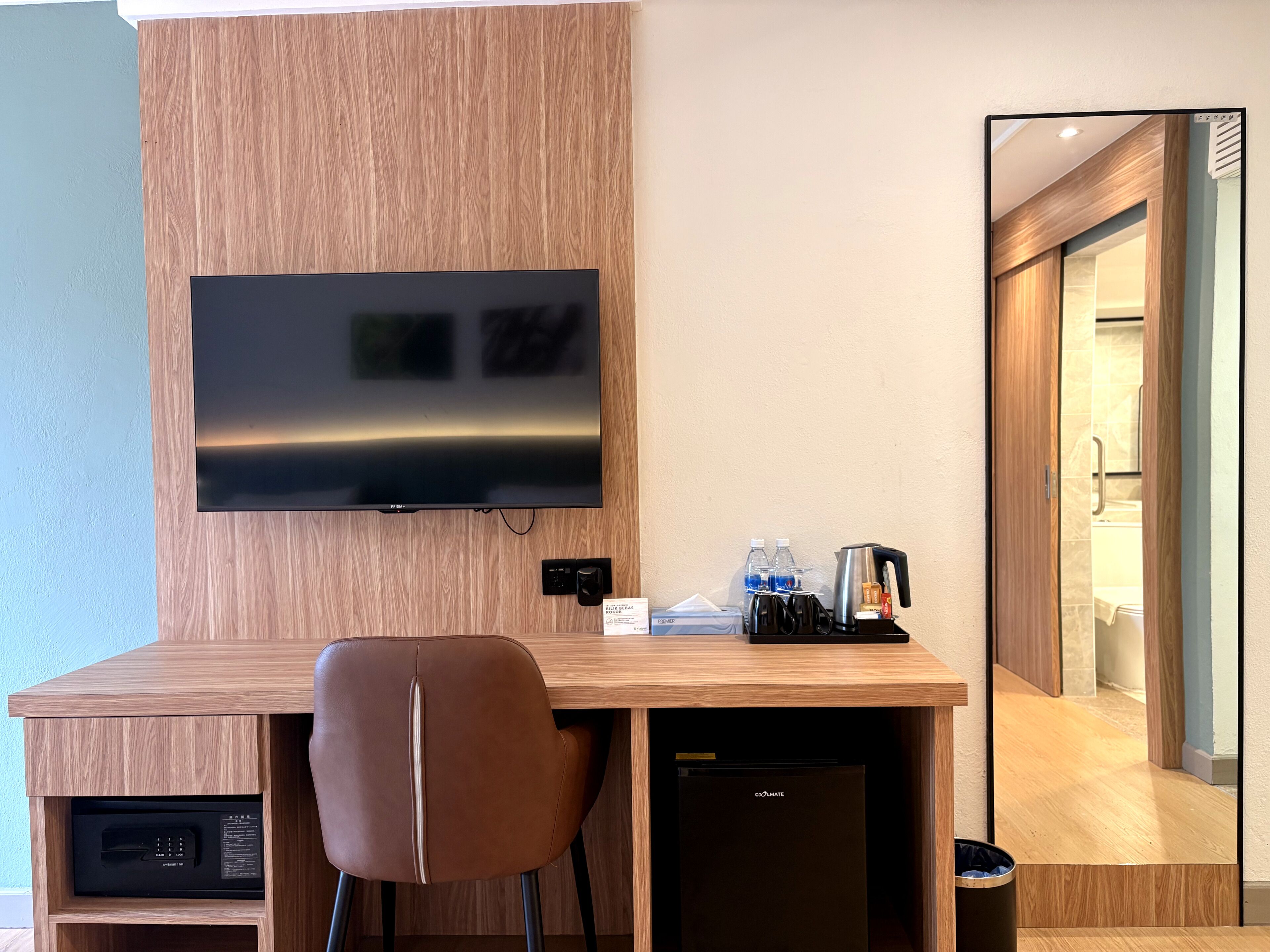 A modern hotel room with a wooden desk, a flat-screen TV, a brown leather chair, a mini-fridge, and a coffee machine.