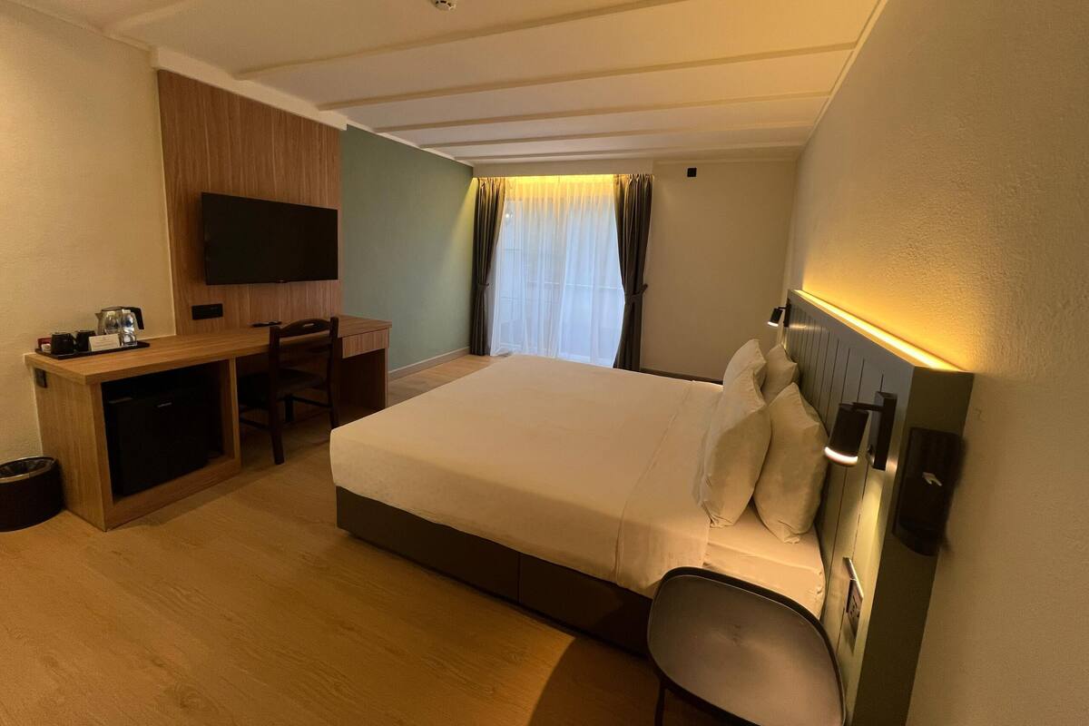 A hotel room with a bed, a desk with a chair, a television mounted on the wall, and a window with curtains.