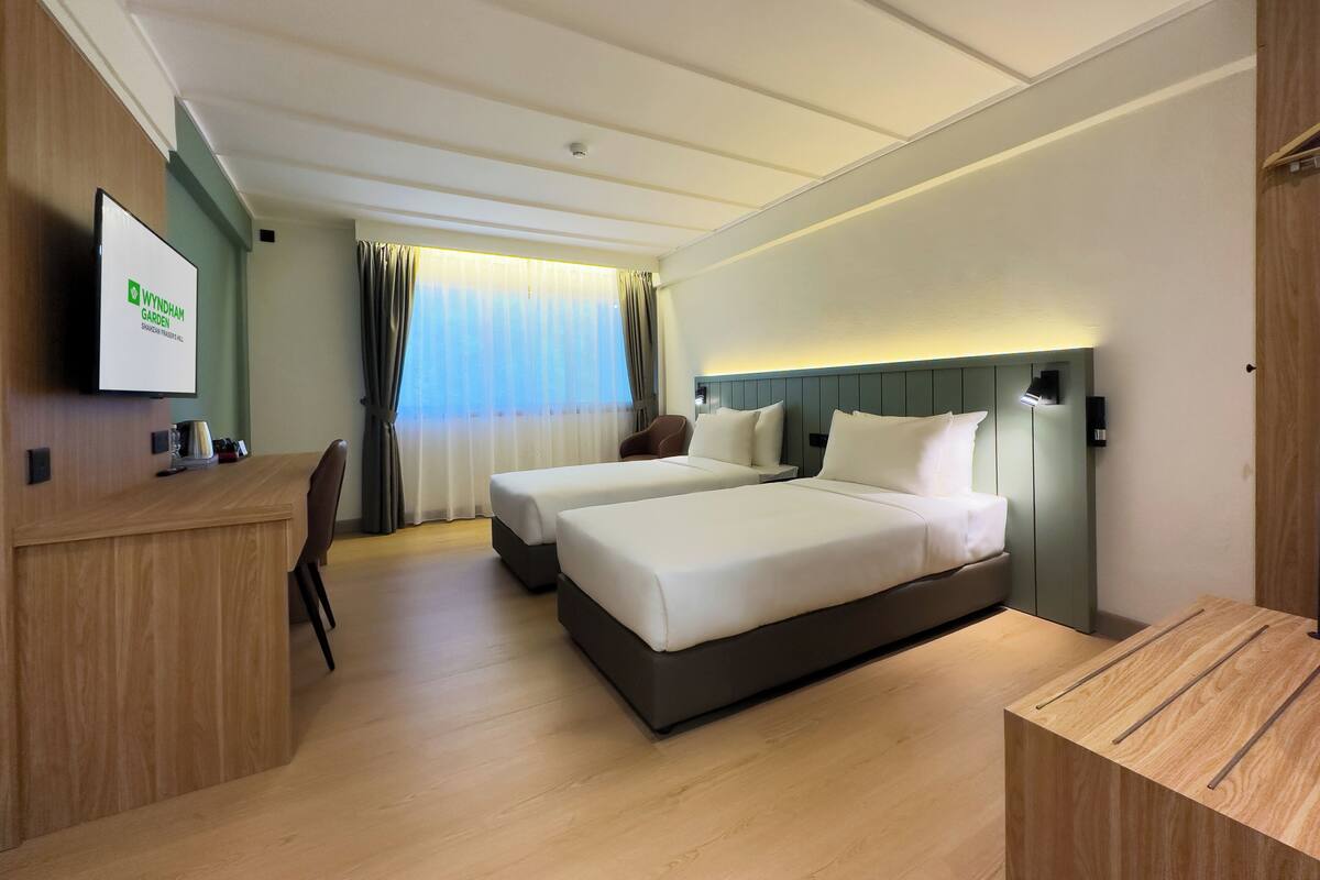 A hotel room with two beds, a desk, a chair, a TV, and a window with curtains.