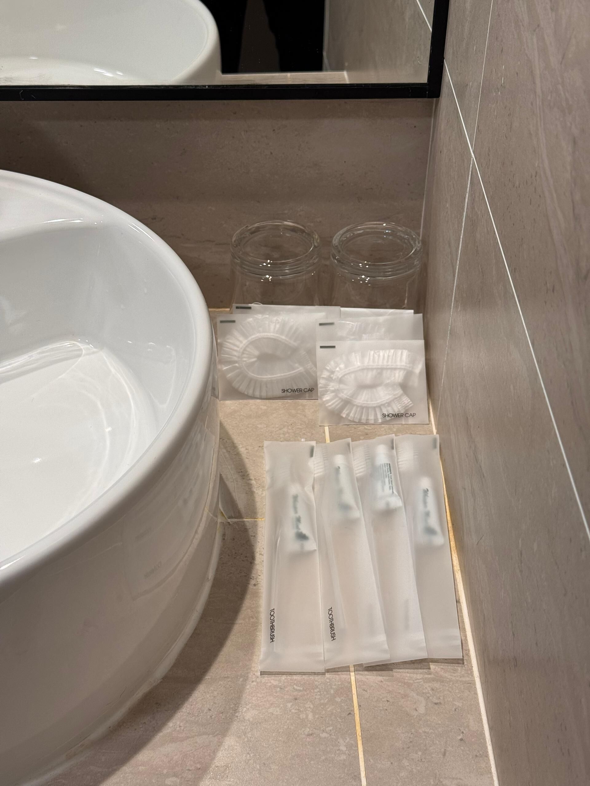 A bathroom with a large white sink, two clear glasses, and several white shower caps and toothbrushes neatly arranged on the floor.