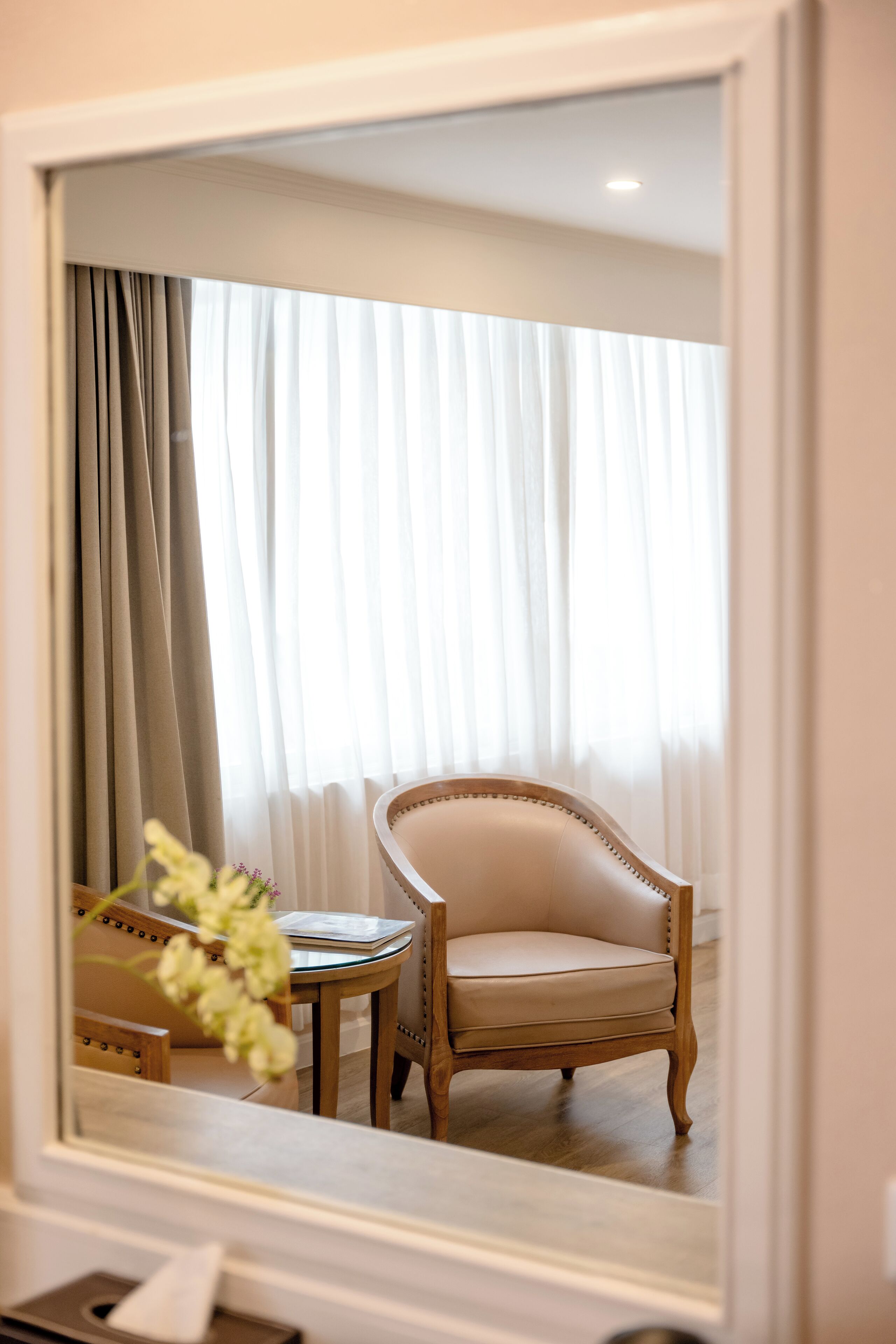 Quarto com um espelho grande refletindo uma cadeira, uma mesa pequena e uma janela com cortinas leves.
