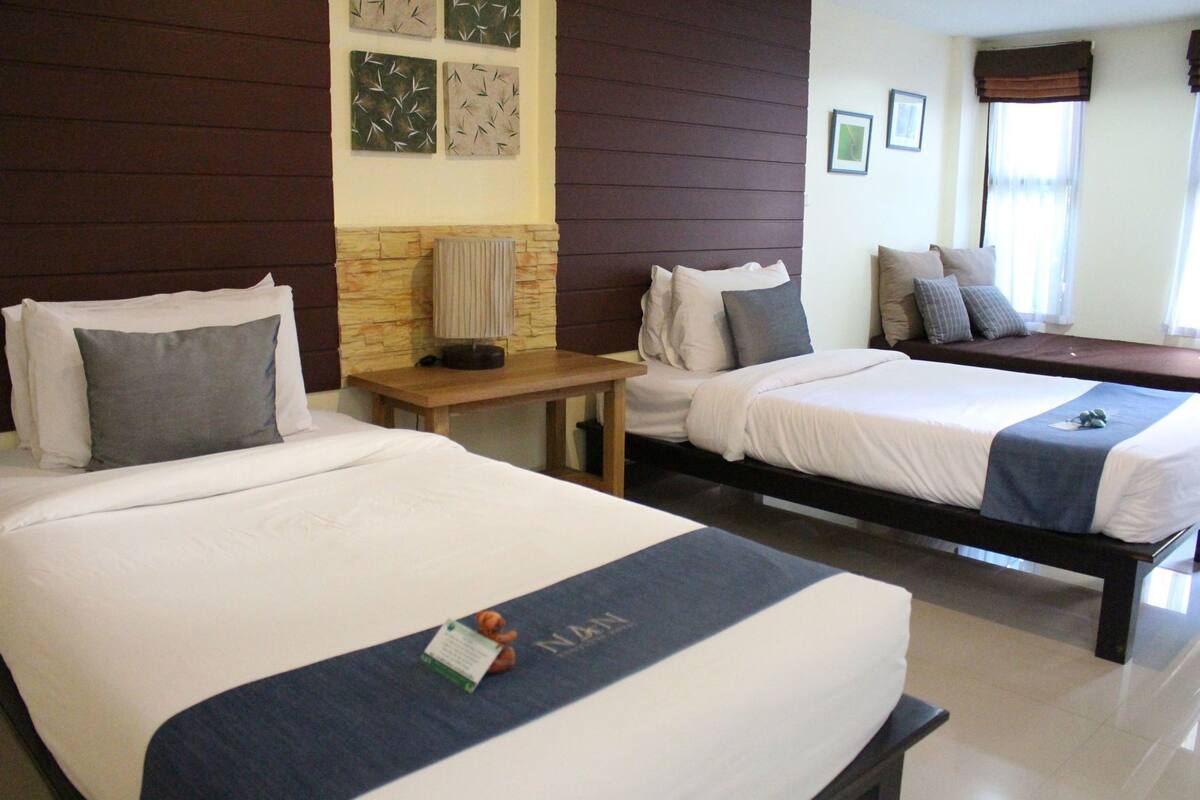A hotel room with two beds, a wooden table, a radiator, and a window with curtains.