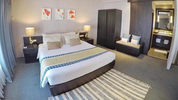 Habitación de hotel con una cama grande, dos mesitas de noche, un armario, un baño con espejo y una pequeña zona de estar.